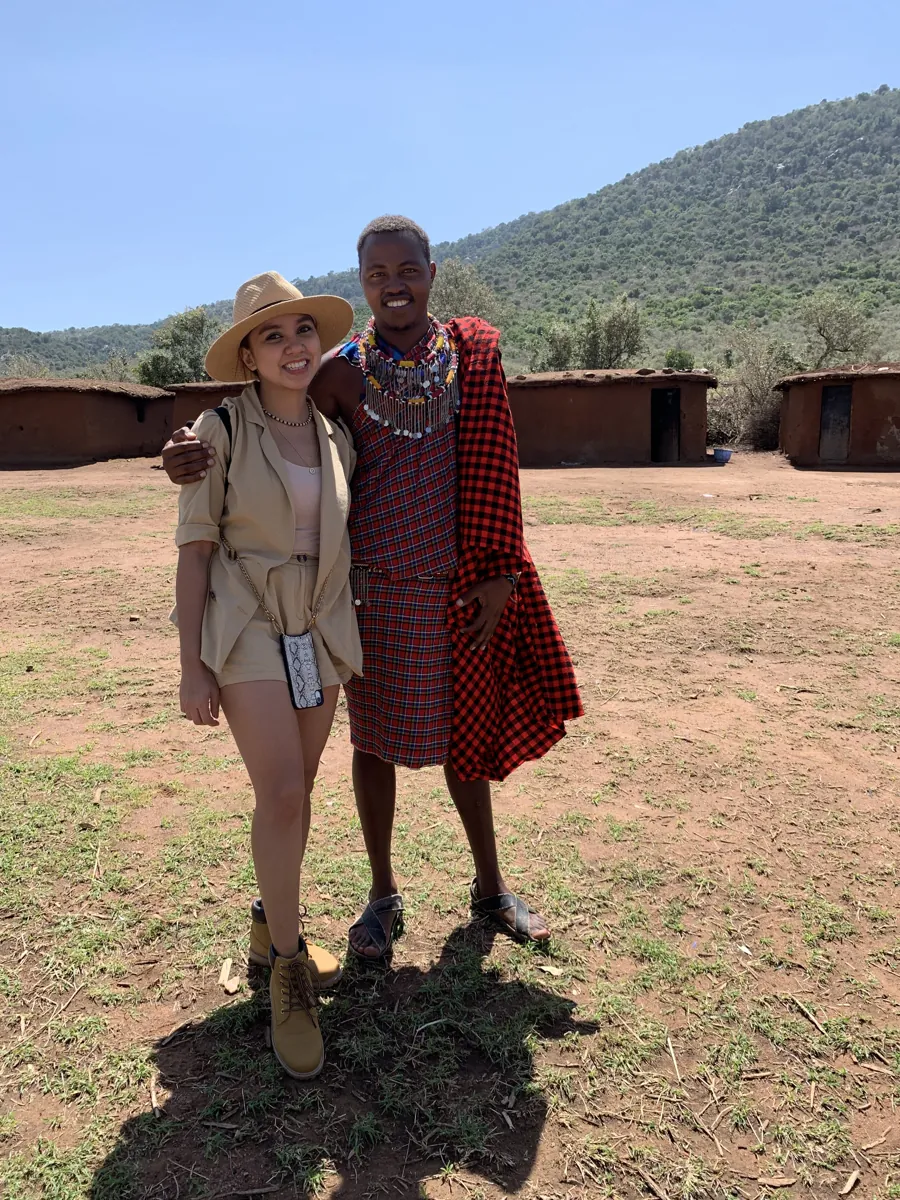 Dee with a Masai man in a red shuka in front of the village