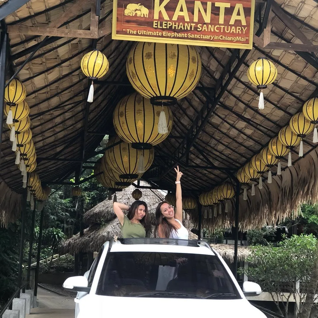 Dee and Kimy waving from a car sunroof under yellow lanterns at the Kanta sign