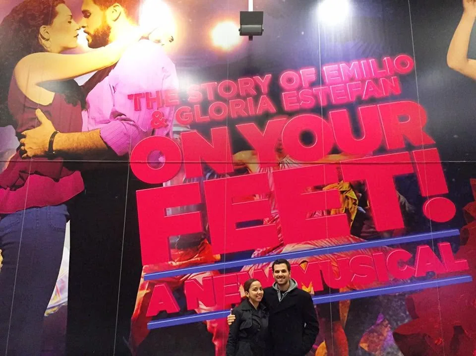 Dee and her husband in front of a giant On Your Feet! poster