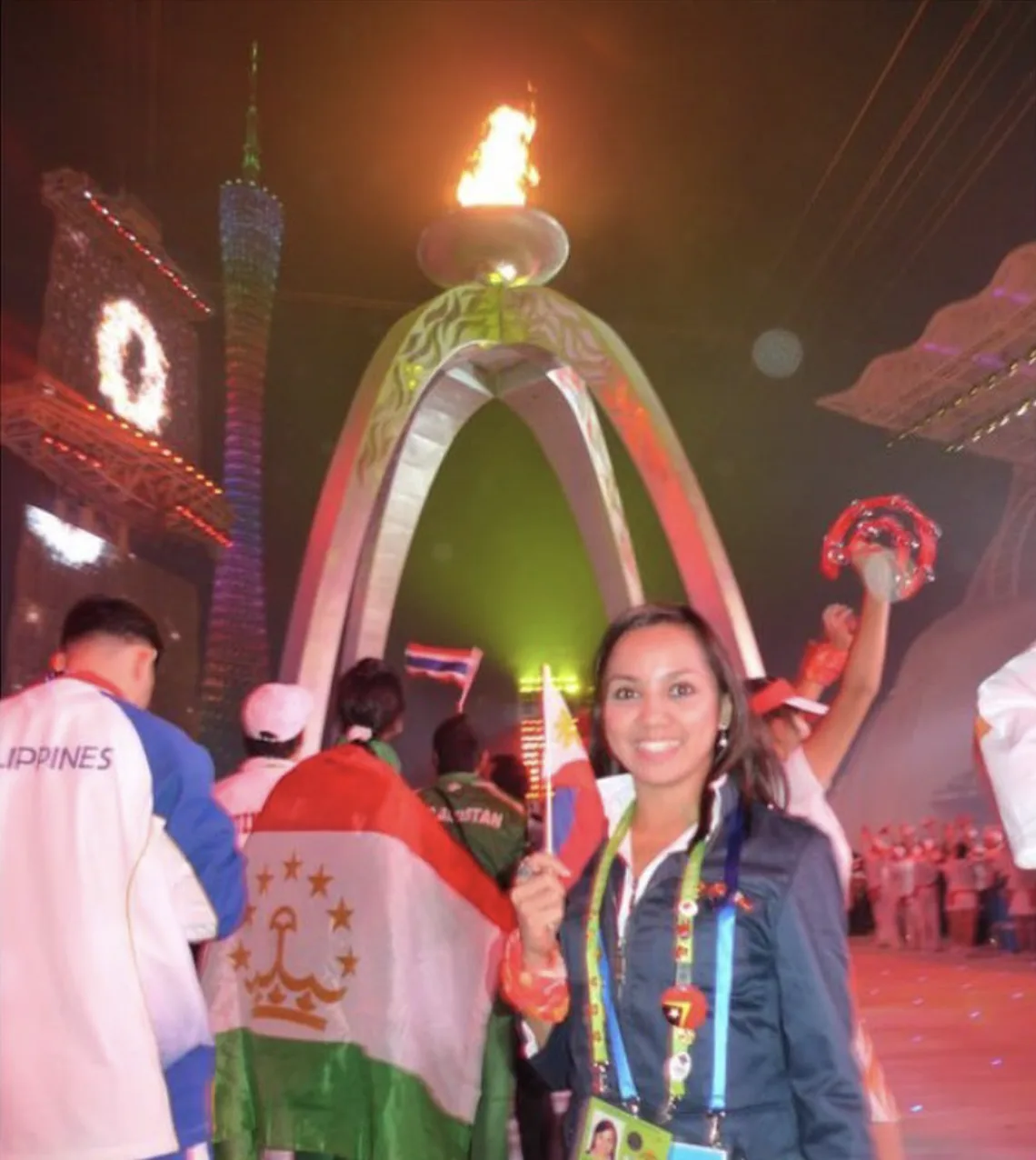 Dee smiling with a small Philippine flag in front of a lit cauldron and a crowd of athletes
