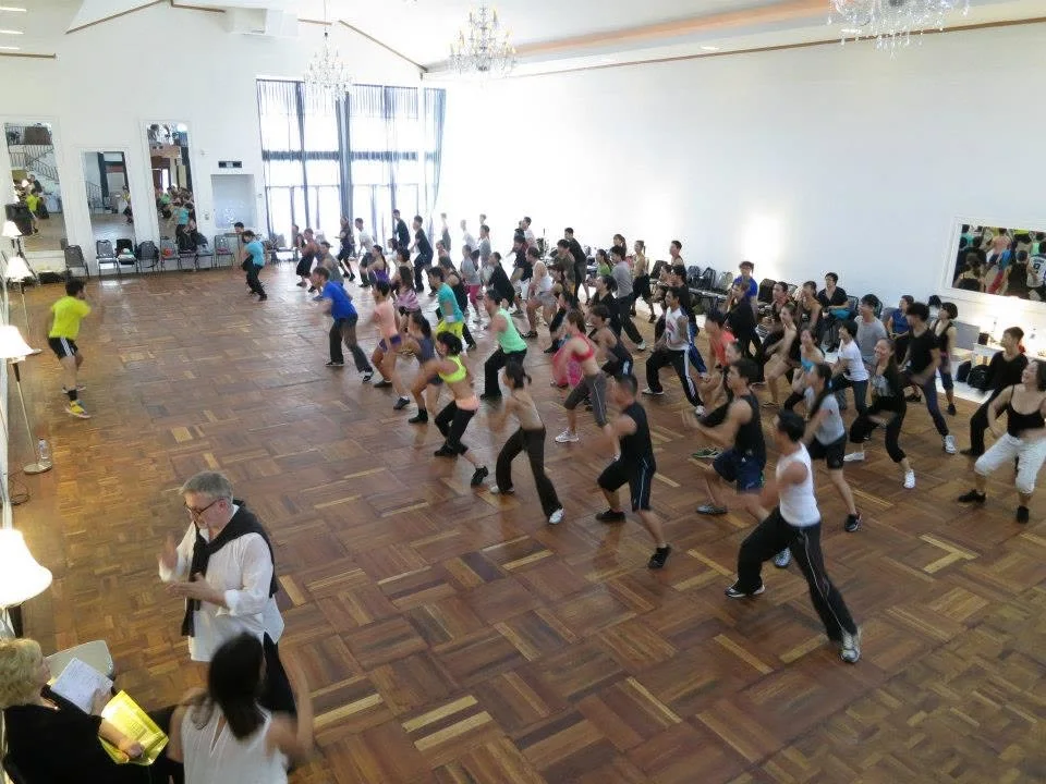 A large group class of dancers lined up across a wide ballroom