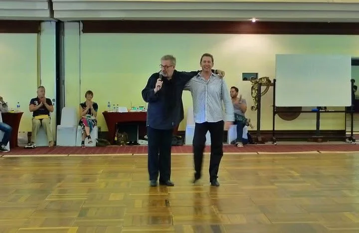 Two dance coaches demonstrating together on a studio floor while dancers watch from the side