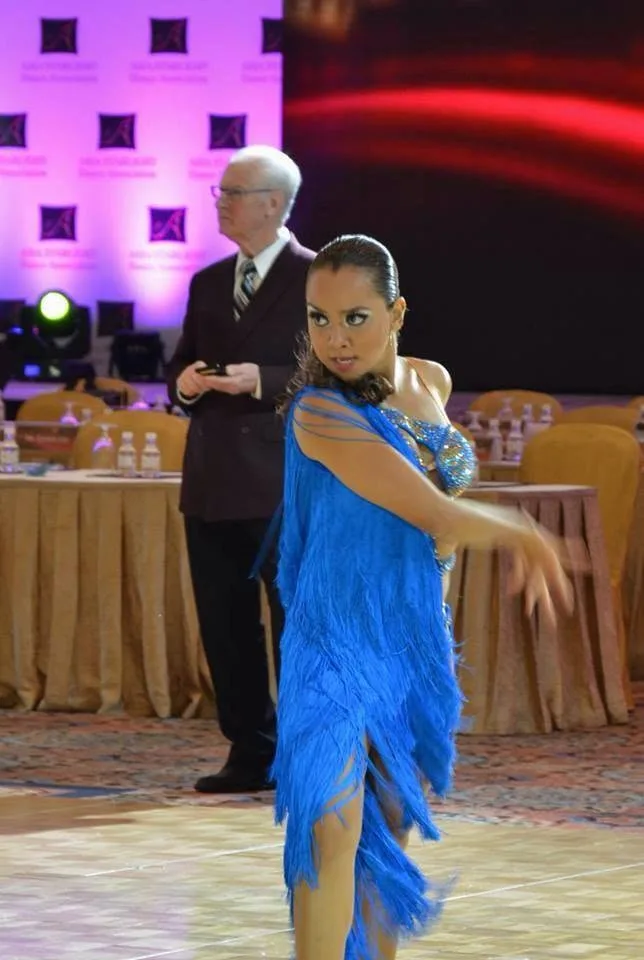 Dee mid-turn in a blue fringed Latin dress at an international competition, an adjudicator behind her